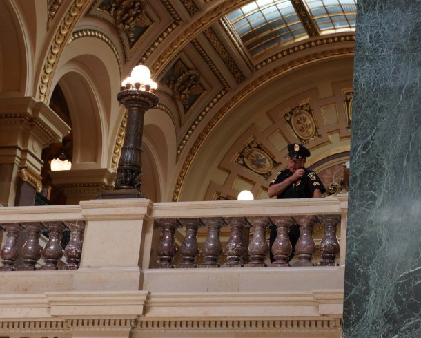 WI Capitol Police Capt Lonergan  speaks into his shoulder communications devive as he directs the arrests of peaceful singers from a second floor balcony. Photo courtesy Leslie Peterson